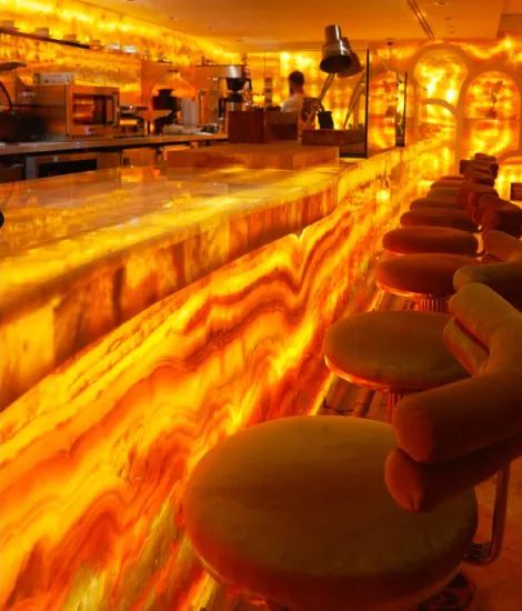 A stylish bar with a glowing onyx countertop, plush stools, and shelves lined with various liquor bottles. Warm, ambient lighting enhances the atmosphere.